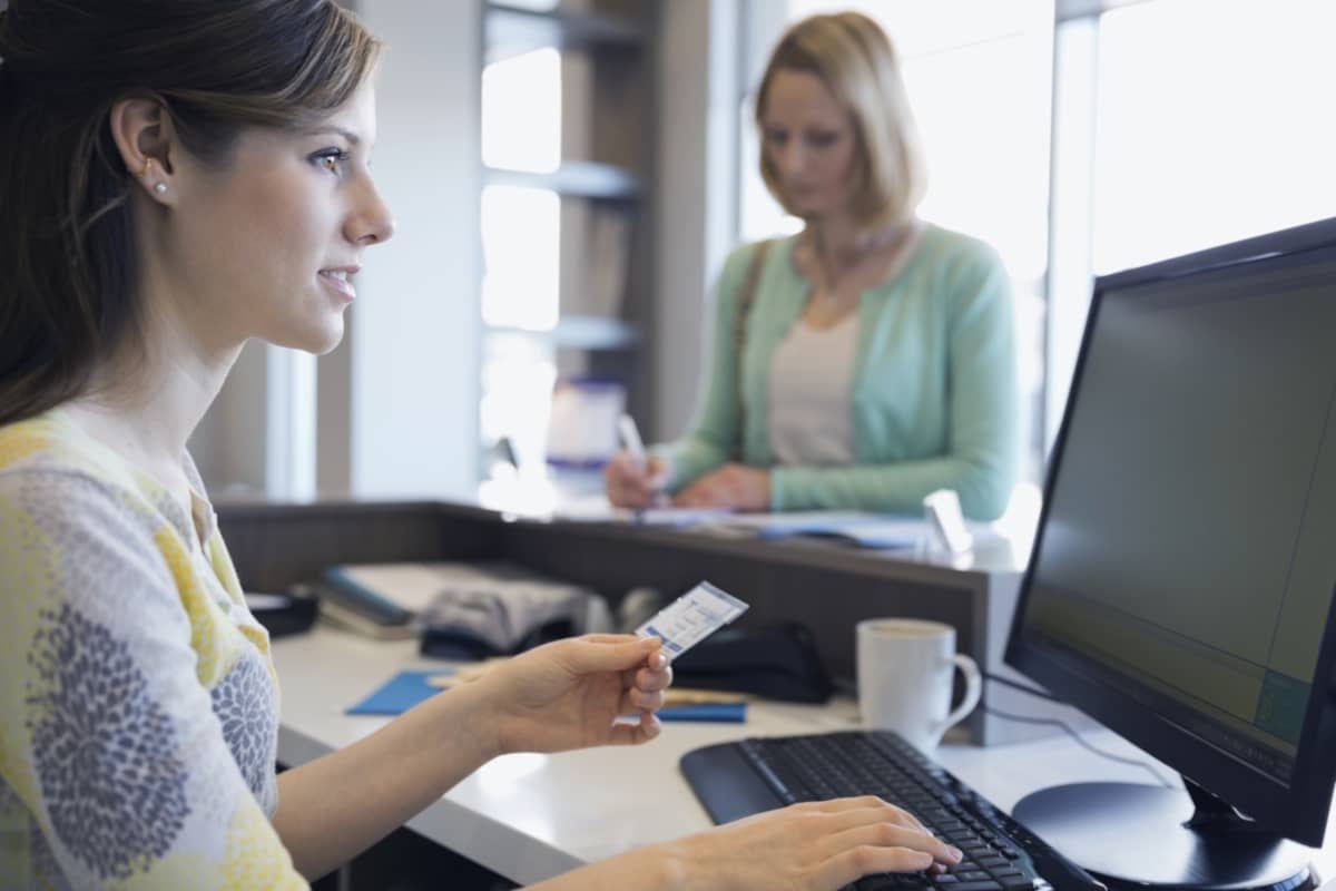 Medical receptionist assisting a patient at a clinical front desk.
